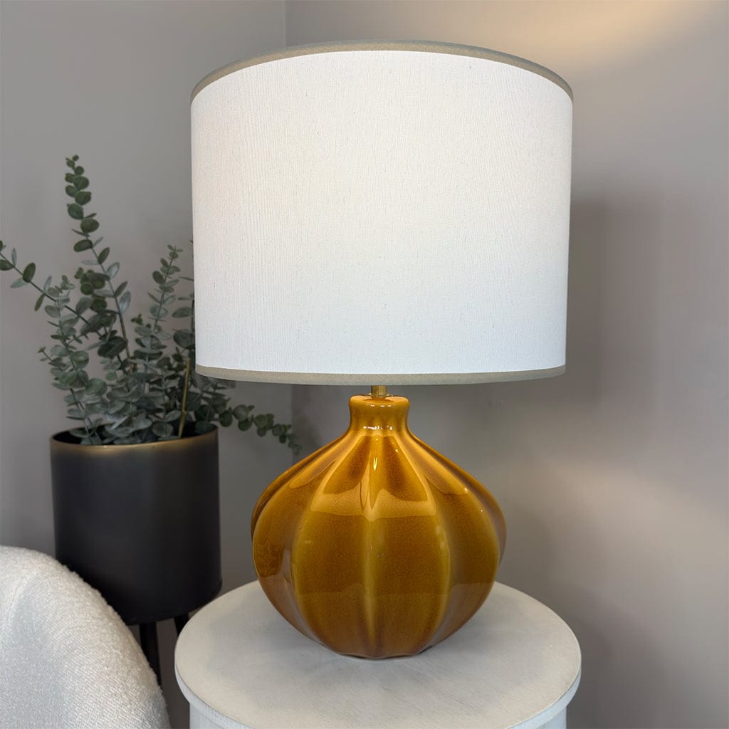 Yellow table lamp with a white shade on a round white table, next to a plant against a gray wall.
