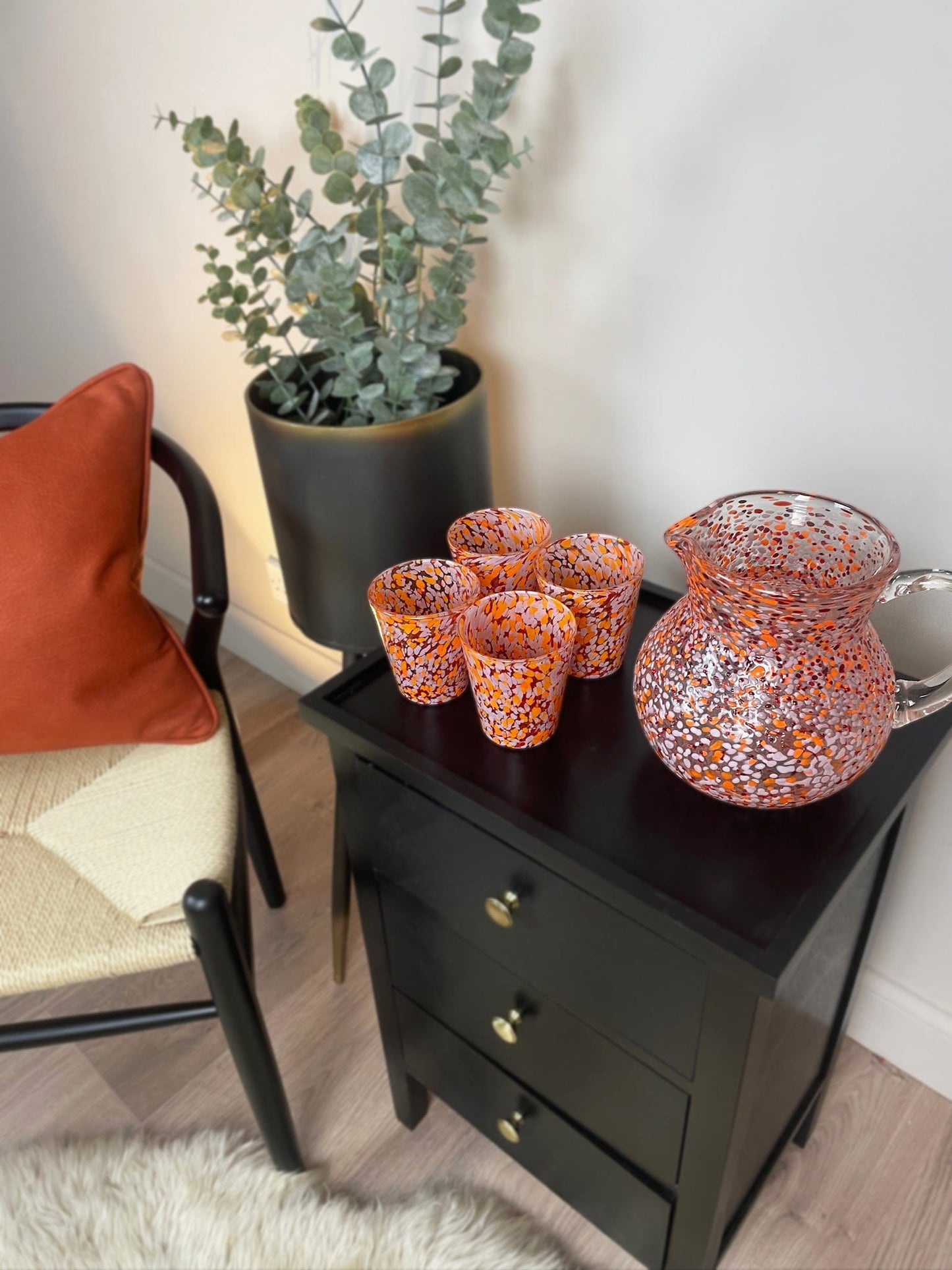 Decorative glass pitcher and cups on a black side table next to a chair with a red pillow.