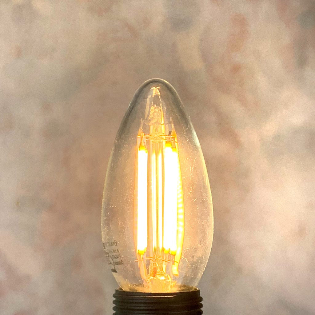 An E14 clear candle non-dimmable LED bulb with visible filaments inside, placed against a textured background, lit