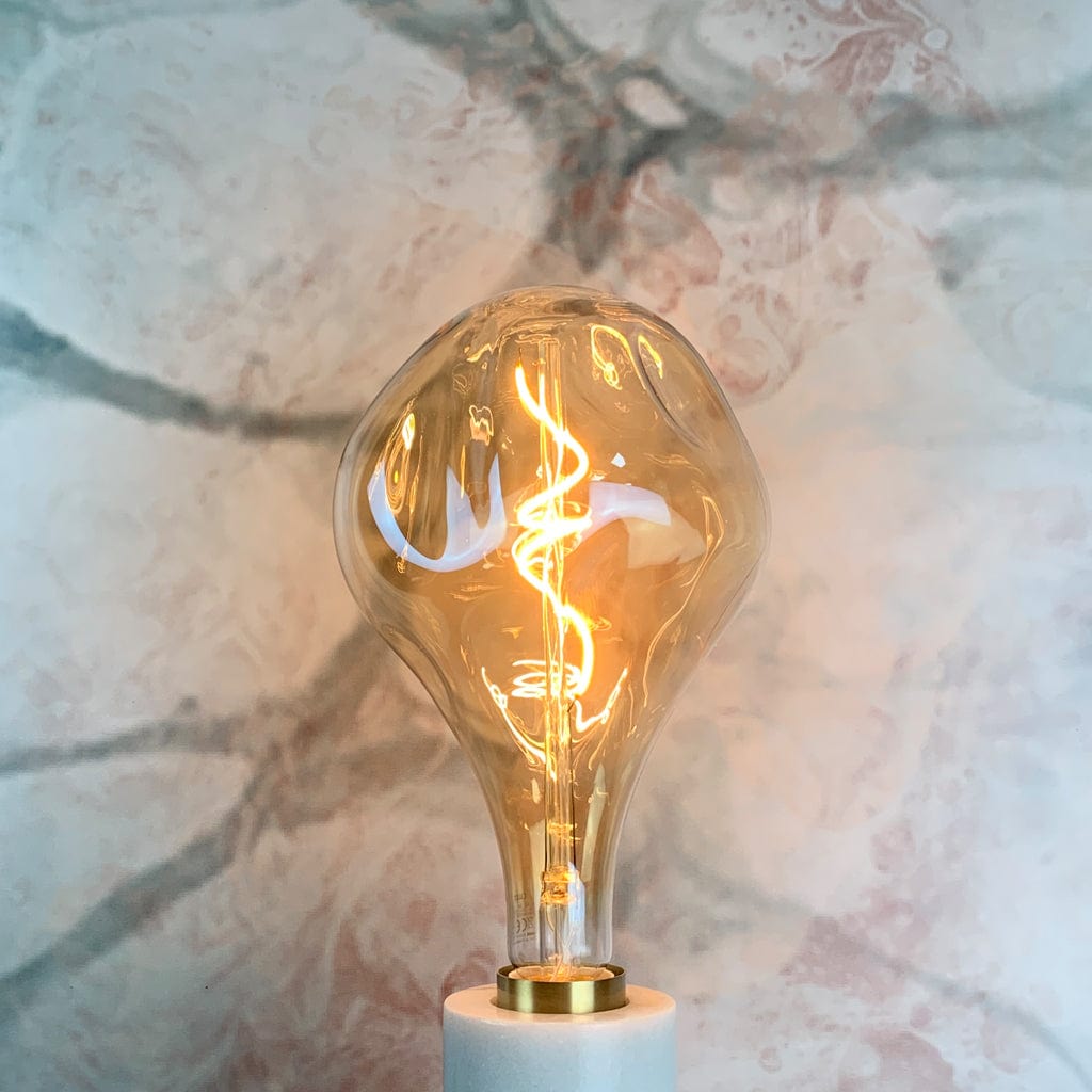 An organically shaped, gold tinted glass LED bulb with visible filament inside, in a lamp holder and lit