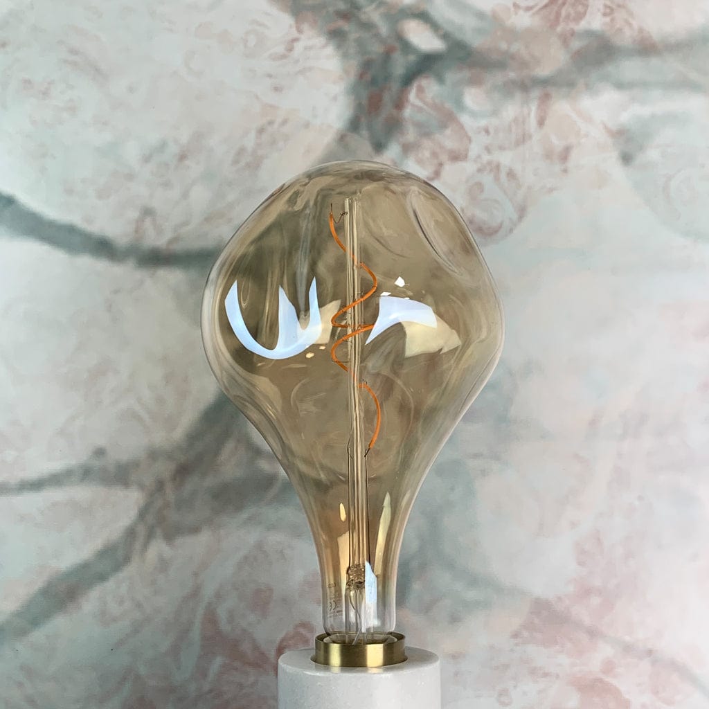 An organically shaped, gold tinted glass LED bulb with visible filament inside, in a lamp holder against a marble background