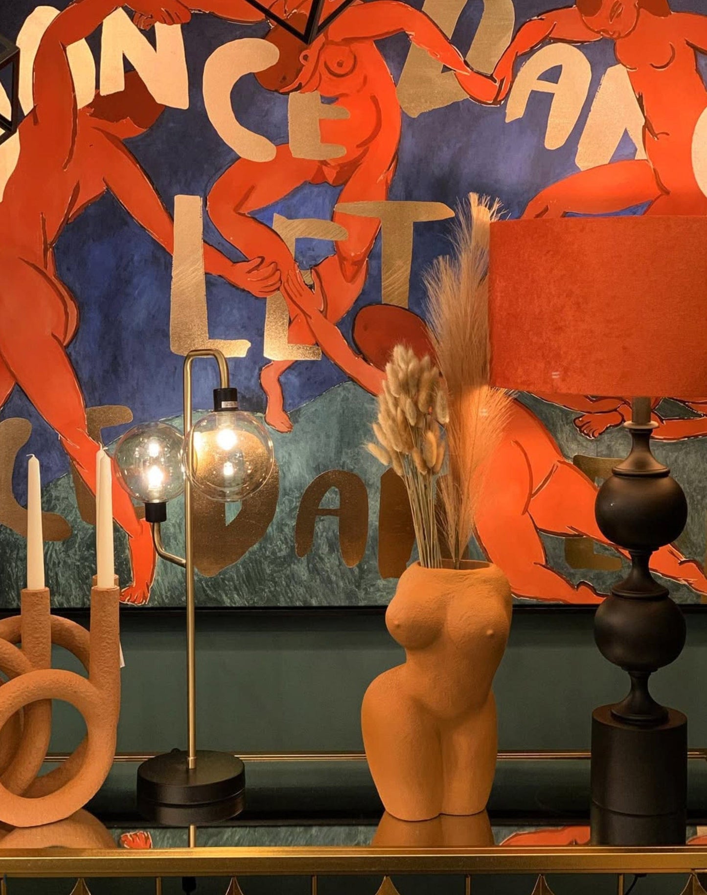 Decorative setting with lamp, vase, and candles against a colorful abstract wall.