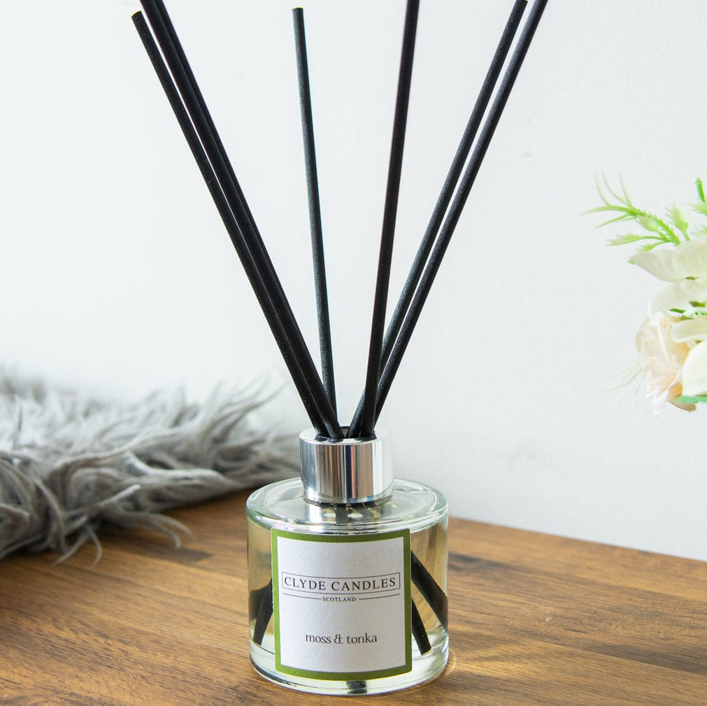 Diffuser with black sticks on a wooden surface with flowers and a blanket in the background