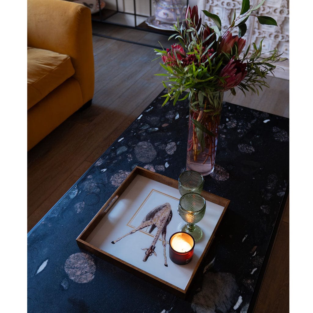 A square giraffe tray with gold tones, placed on a table with a floral pattern, serving as a platter for a candle and a flower vase.