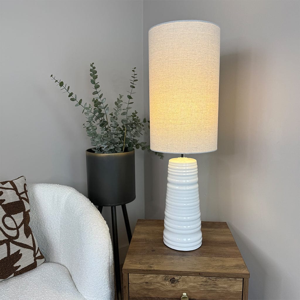 White textured table lamp on a wooden side table with a plant and cushion in the background.