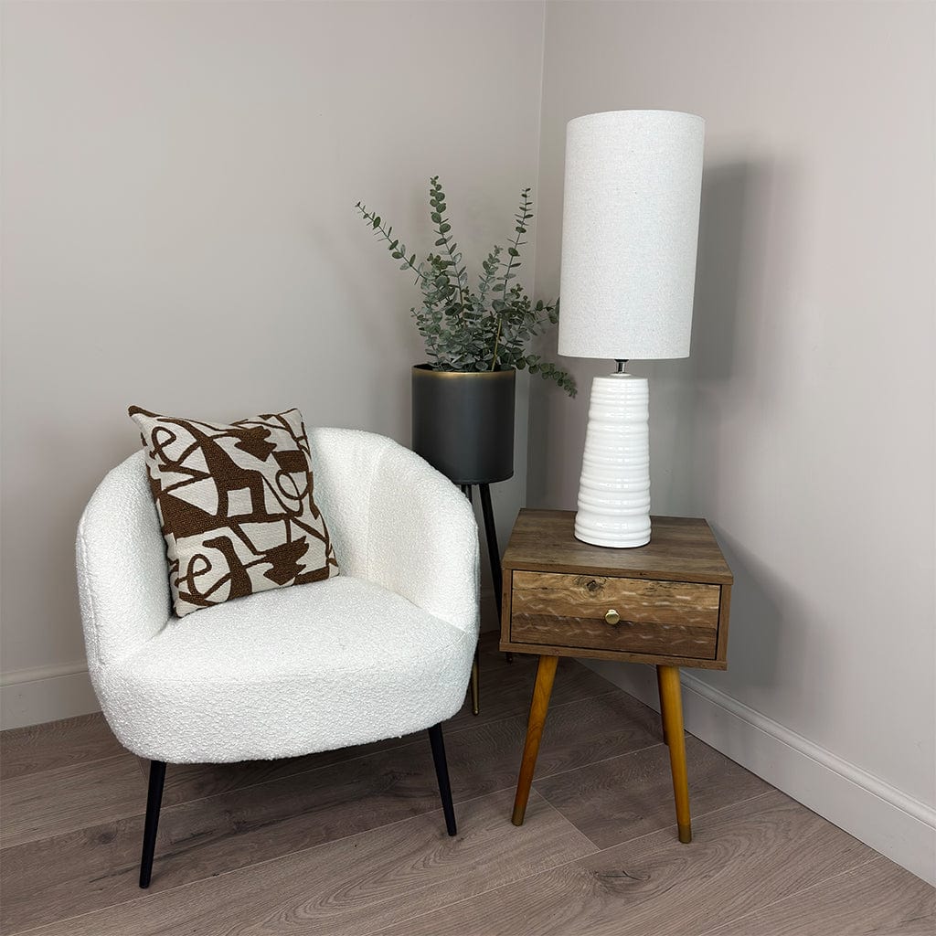 White armchair with a patterned pillow, wooden side table with a lamp, and plant in a room.