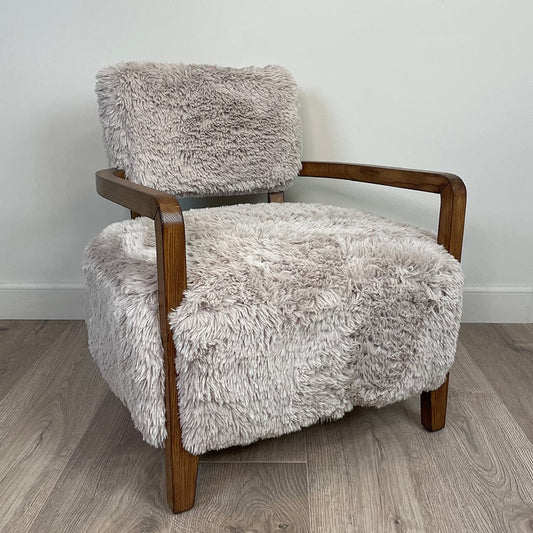 A caramel-colored faux fur armchair with dark ash wood arms, displayed in a cosy indoor setting.