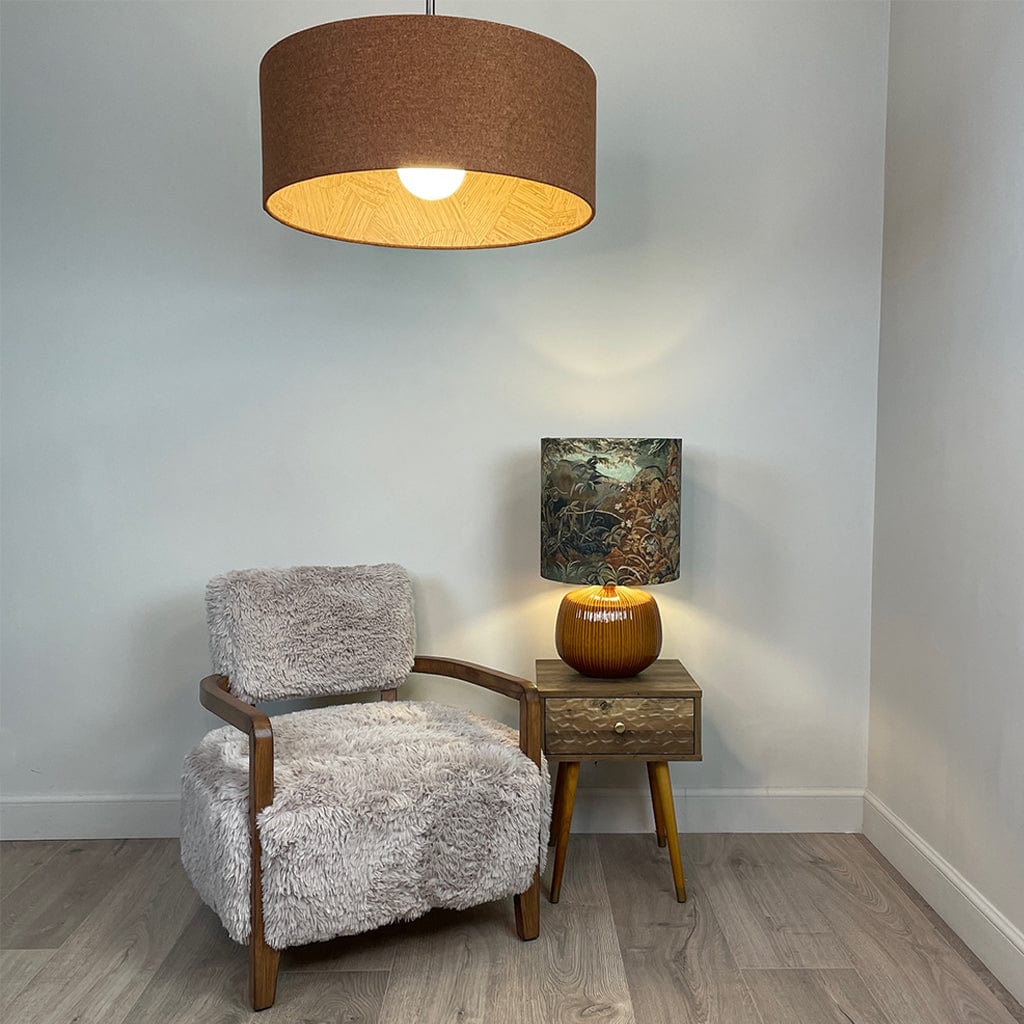 A caramel-colored faux fur armchair with dark ash wood arms, displayed in a cozy indoor setting featuring a small wooden side table, an ochre lamp base with a forest shade and a large orange shade overhead