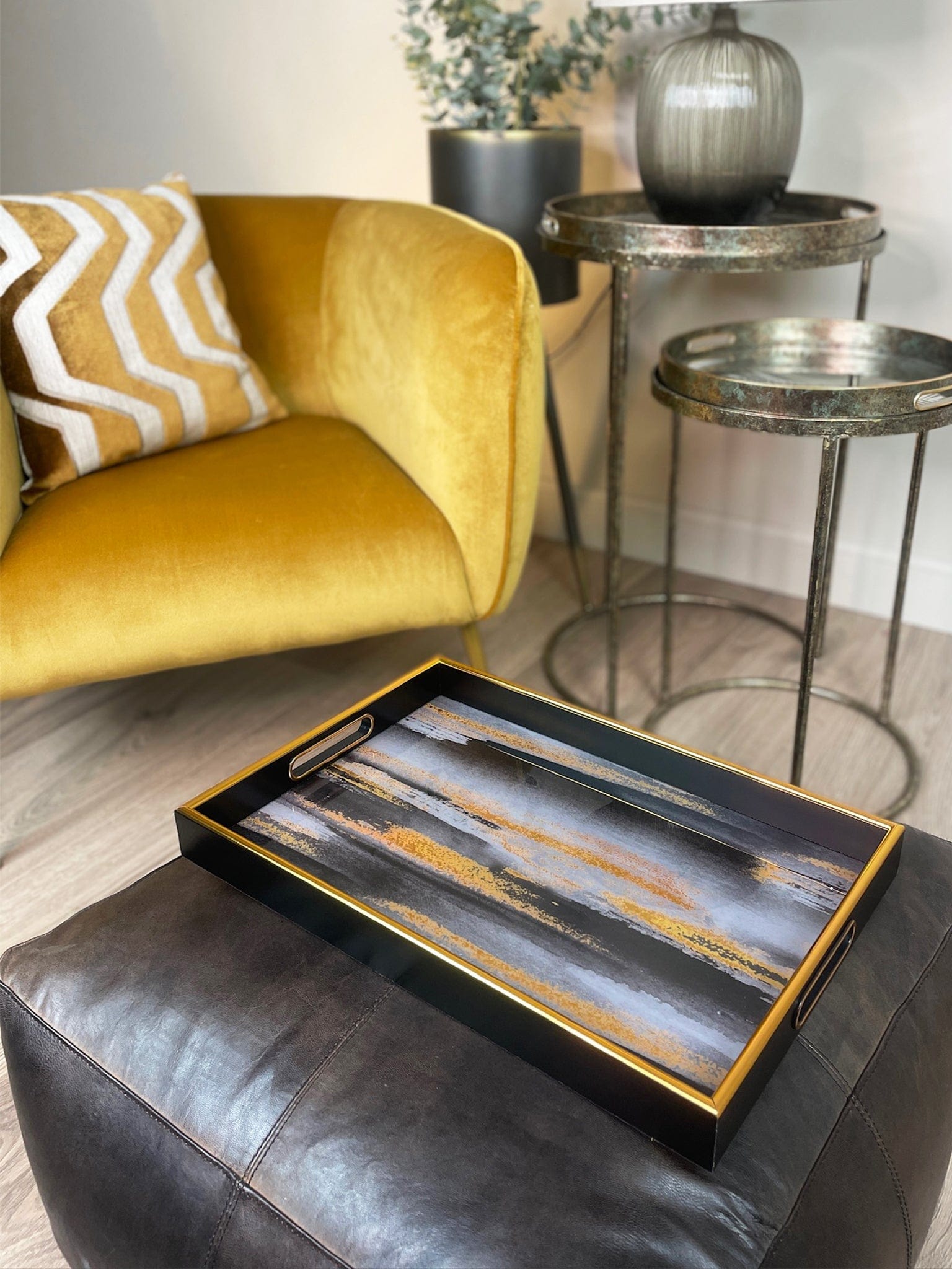 Decorative tray on a black leather ottoman with a yellow armchair and side table in the background.