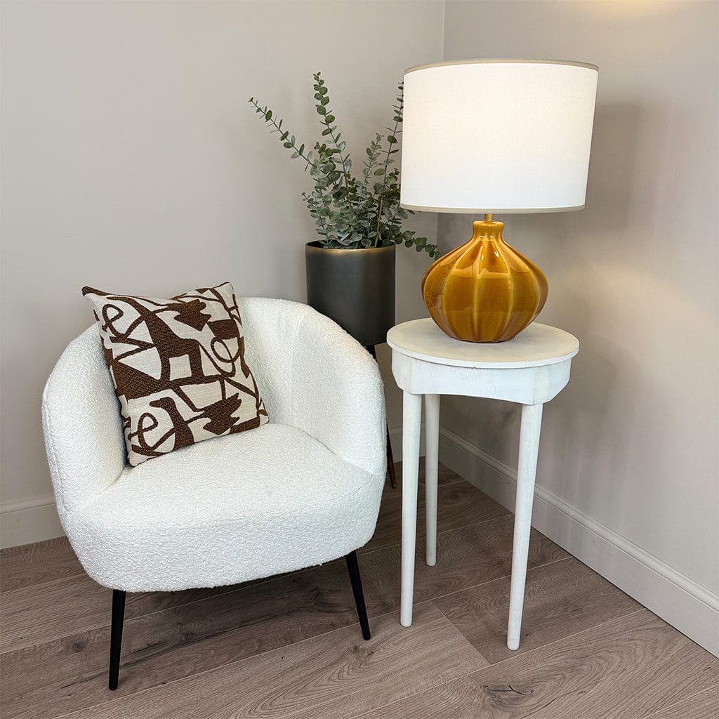 White armchair with a decorative pillow, small round table with a lamp, and plant in a room.