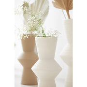 A sleek cream-colored ceramic vase with a geometric design, displayed with other vases in the background.