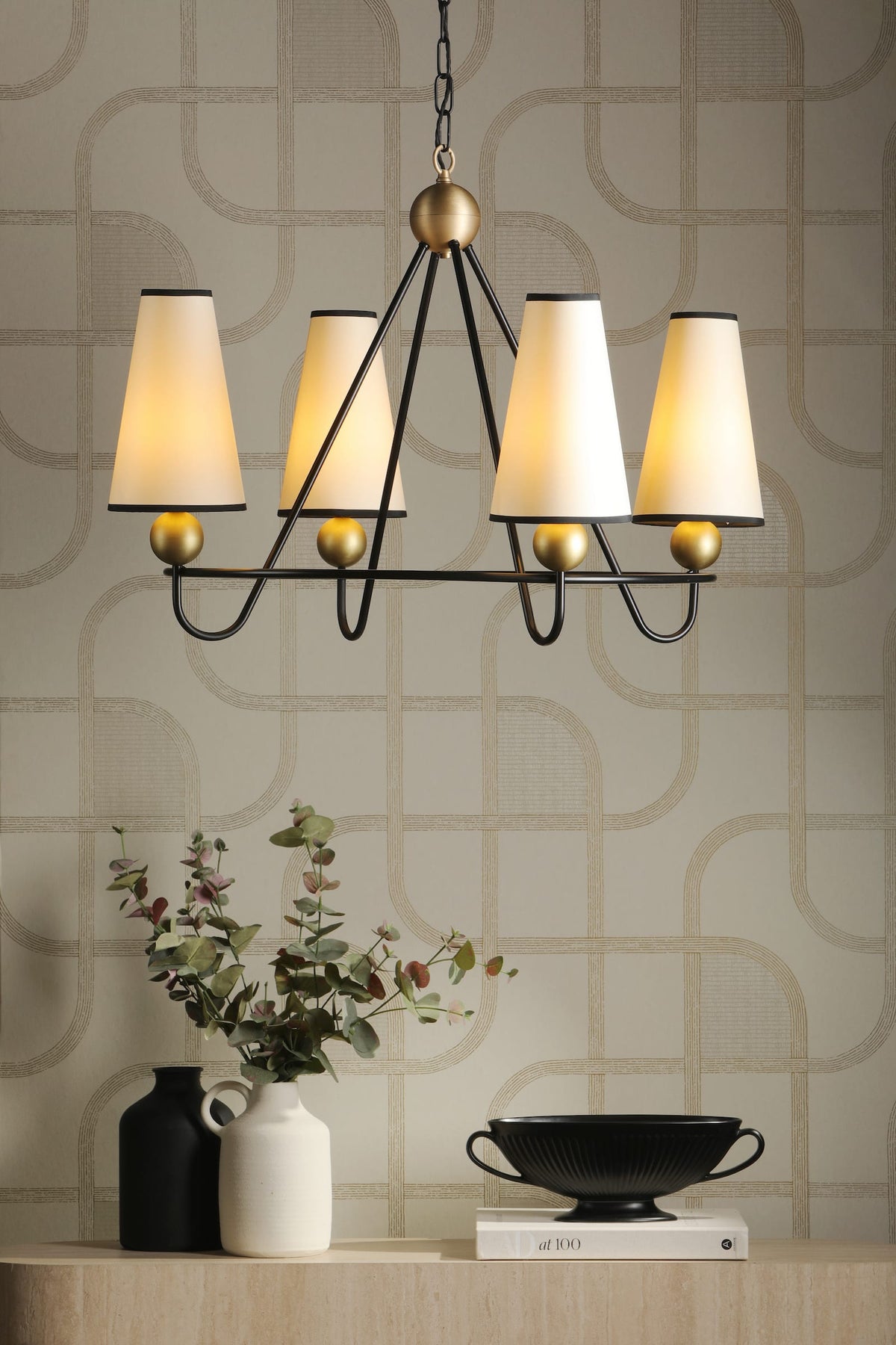 A modern five-light chandelier with a black metal frame, warm gold accents, and tapered cream fabric shades, hanging in a styled room above a console table with vases and a bowl.