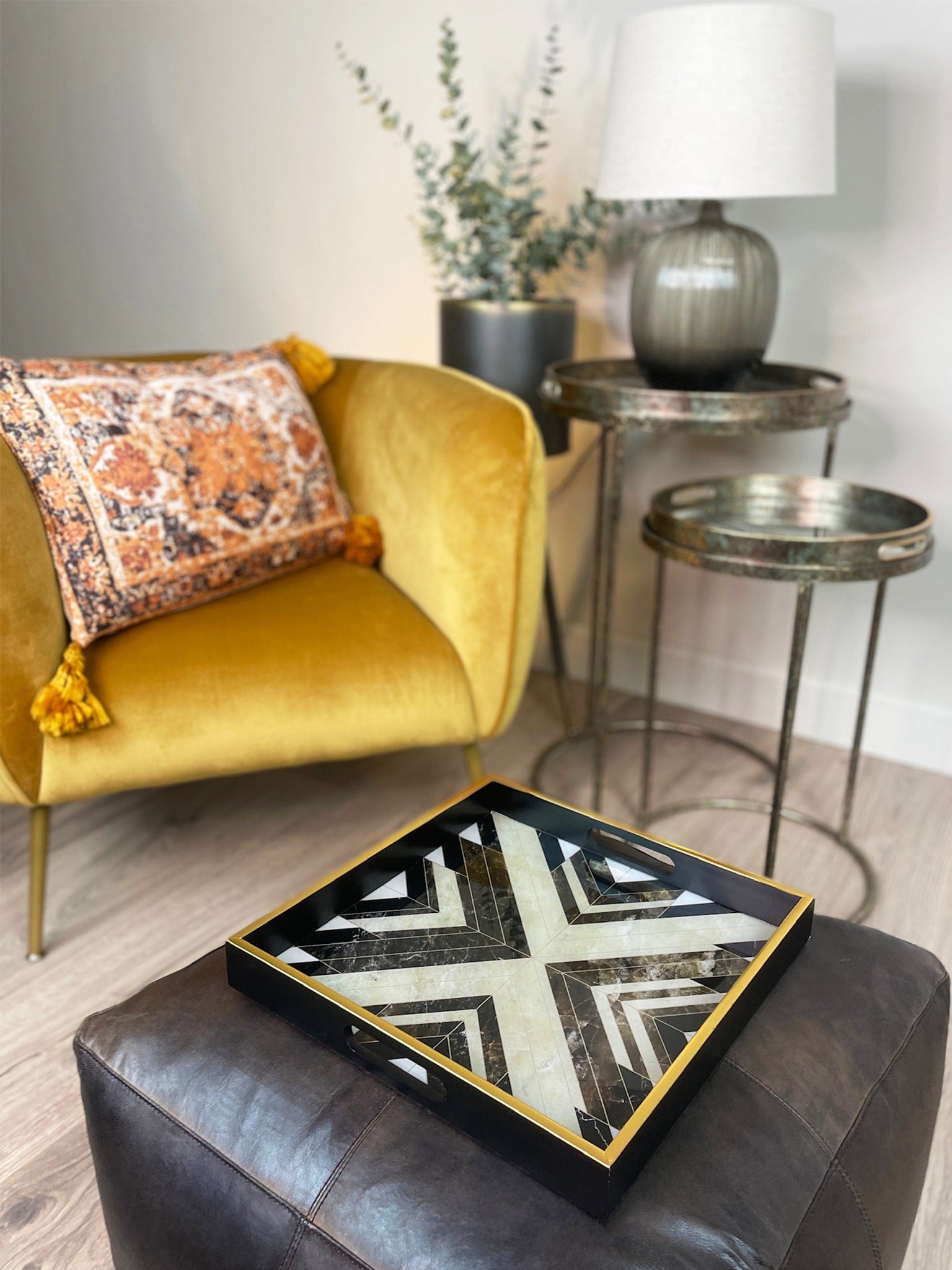 Living room with yellow armchair, patterned pillow, and decorative coffee table. There is a black and gold geometric design on a square tray on a brown leather ottoman