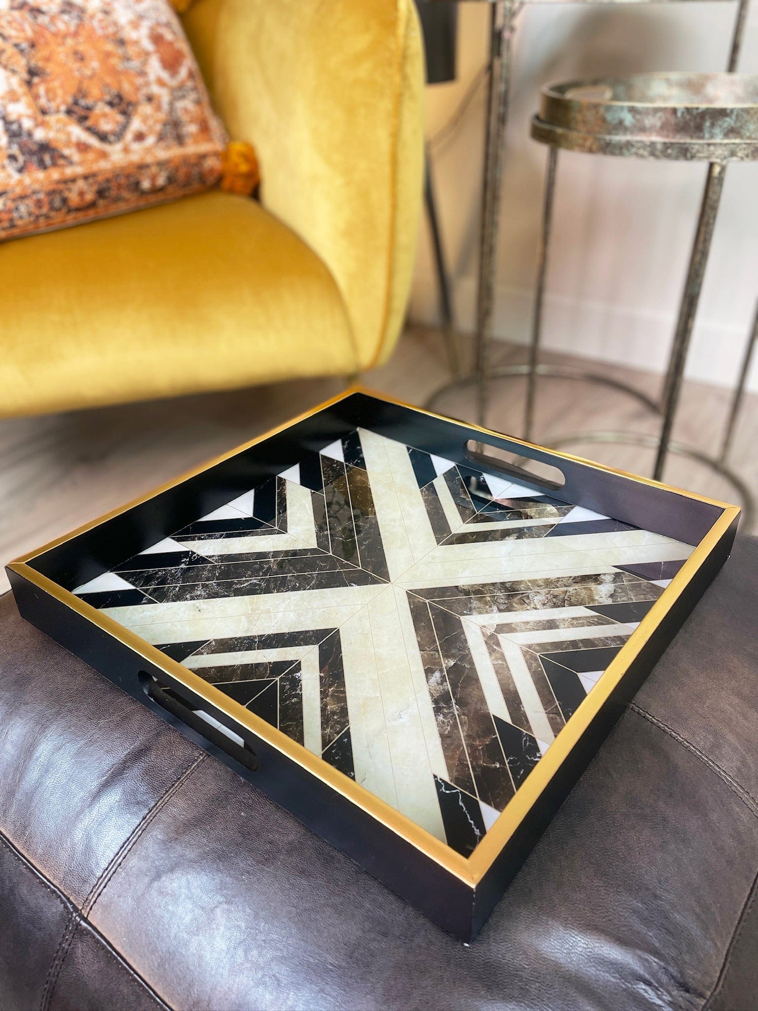 Decorative tray with a black and gold geometric design on a leather ottoman, with a yellow armchair and side table in the background.