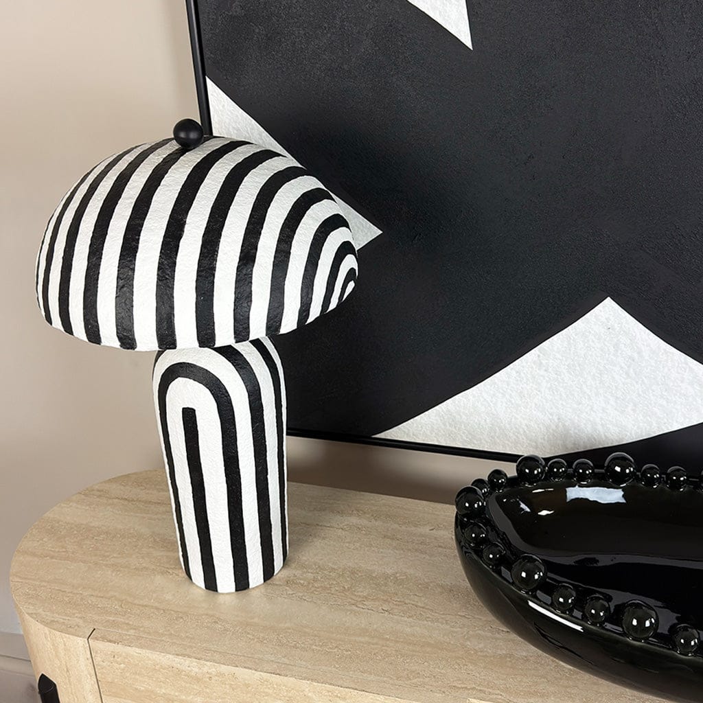 Black and white striped table lamp on a wooden surface with a black and white abstract painting in the background.
