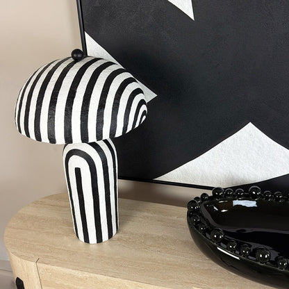 Black and white striped table lamp on a wooden surface with a black and white abstract painting in the background.
