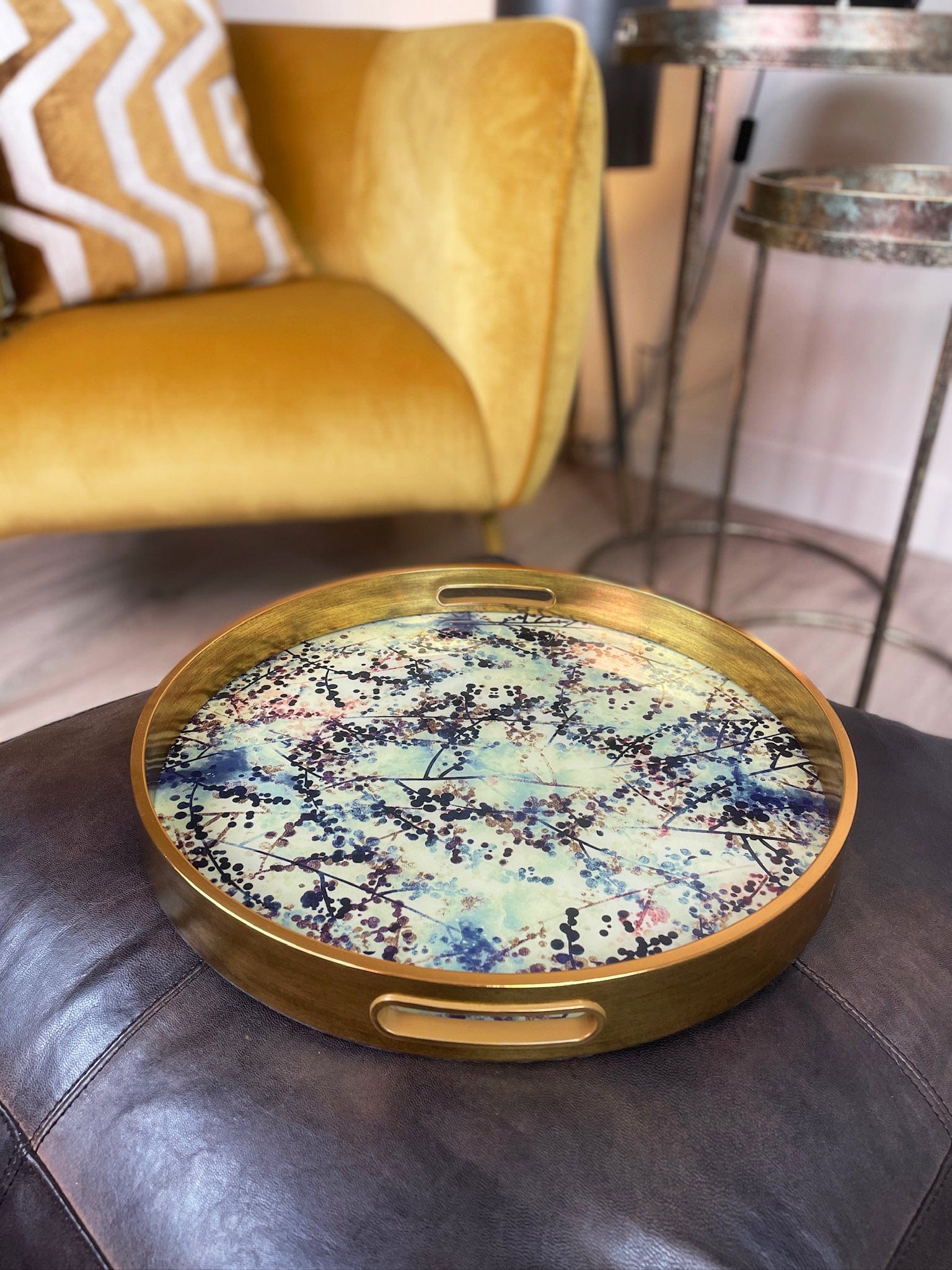 Round decorative tray with artistic design on a dark surface, with a yellow armchair in the background.