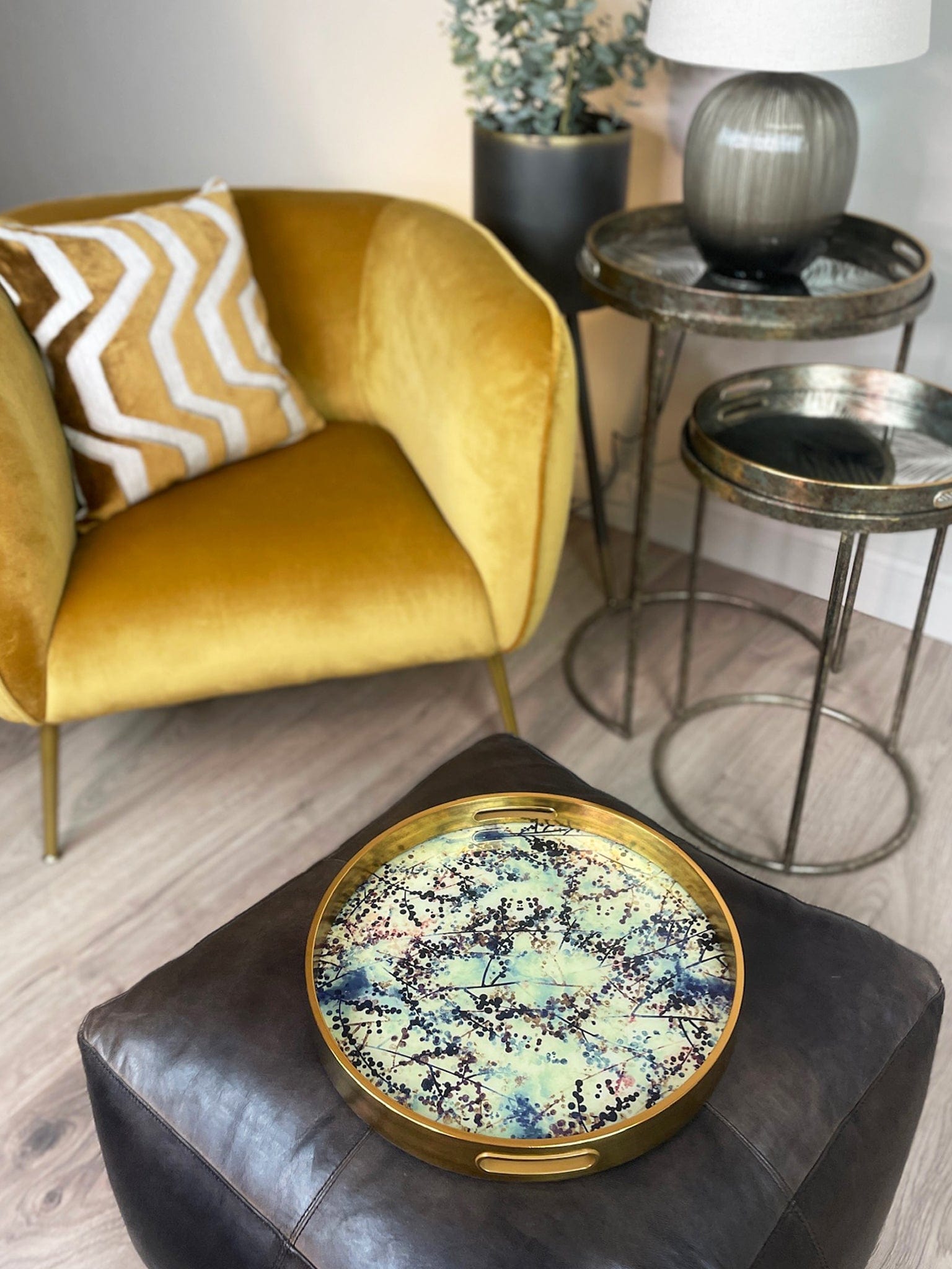 Decorative round tray with gold rim on a black ottoman in a living room setting.