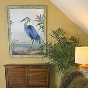 Framed painting of a blue heron on a wall above a wooden cabinet with a plant and lamp in the background.