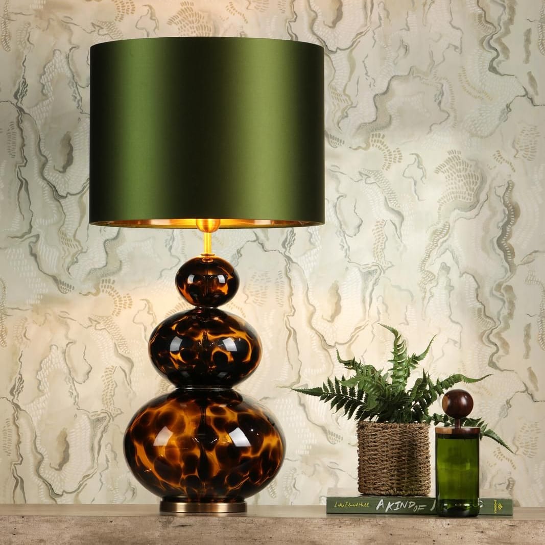 A table lamp with a dark green cylindrical shade and a stacked amber-brown glass base, displayed on a tabletop against a textured beige wall with a small plant and decorative bottle beside it.