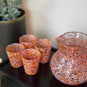 Decorative glass pitcher and cups on a black side table next to a chair with an orange pillow and a plant.