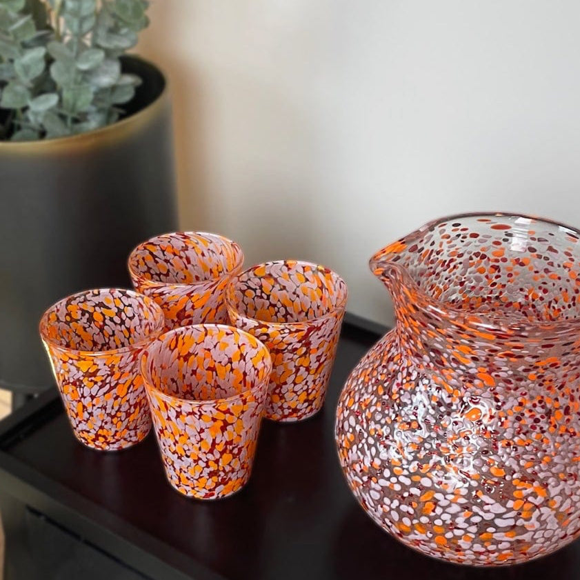 Decorative glass pitcher and cups on a black side table next to a chair with an orange pillow and a plant.