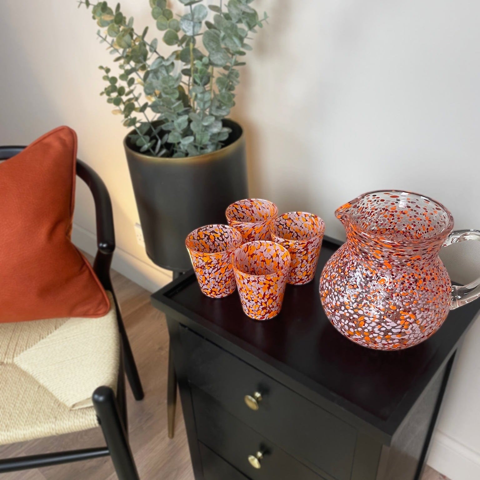 Decorative glass pitcher and cups on a black side table next to a chair with a red pillow.