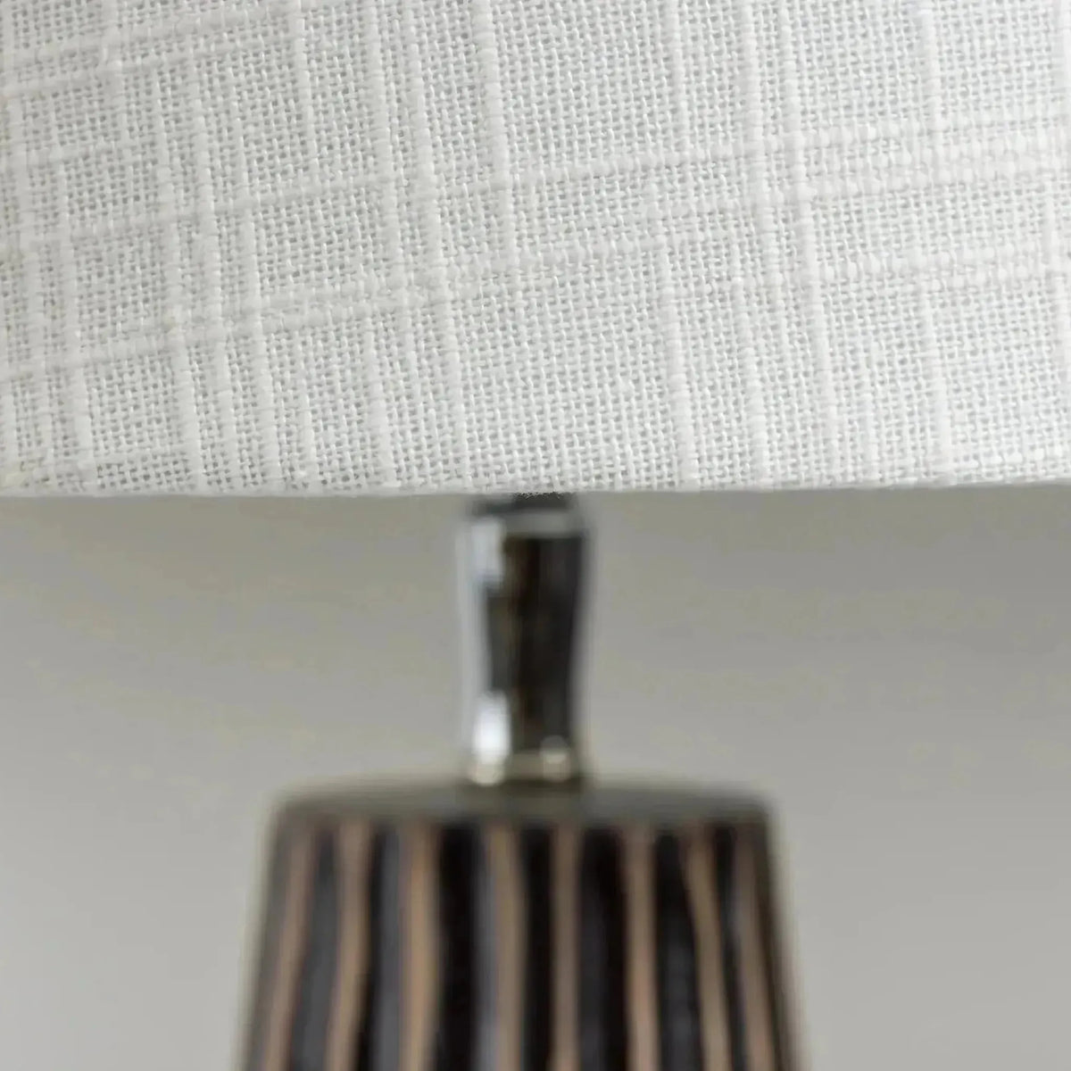 Close-up of a table lamp with a white woven fabric shade and a dark brown vertically striped base.