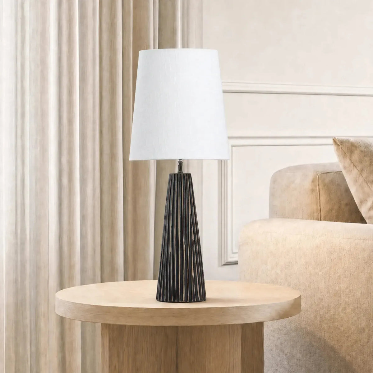 A light oak round end table in a modern living room setting next to a beige sofa, featuring a black ribbed table lamp with a white drum shade.