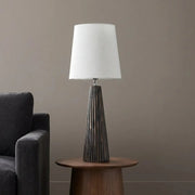 A table lamp with a white drum shade and a dark gray ribbed tapered base, styled on a round wooden side table next to a dark gray armchair against a neutral gray wall.