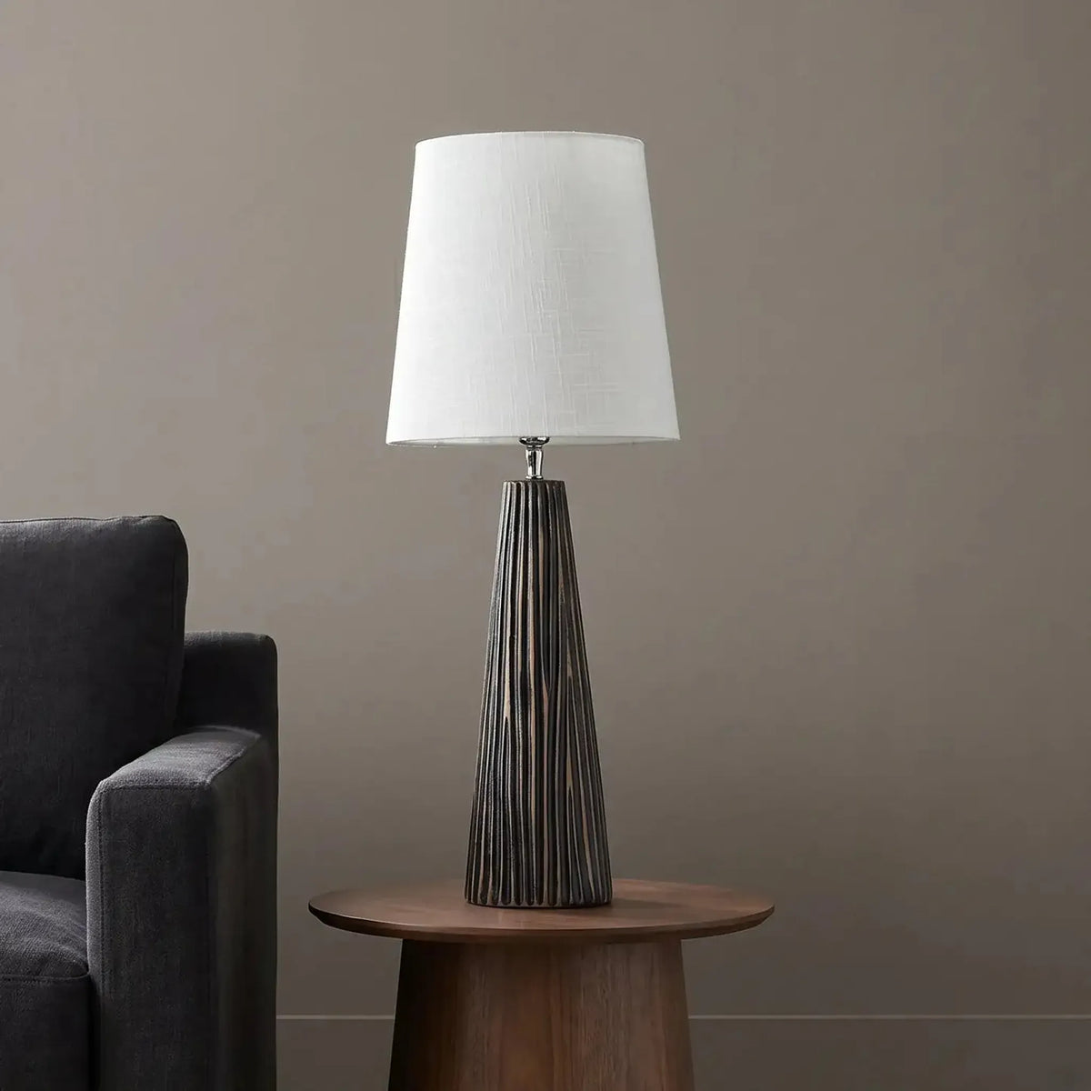 A table lamp with a white drum shade and a dark gray ribbed tapered base, styled on a round wooden side table next to a dark gray armchair against a neutral gray wall.