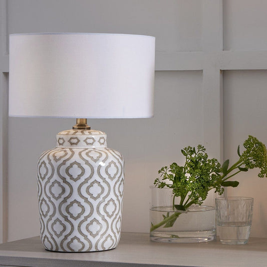 A ceramic table lamp with a taupe and white geometric pattern, featuring a white poly cotton shade, placed on a surface with a plant in the background.
