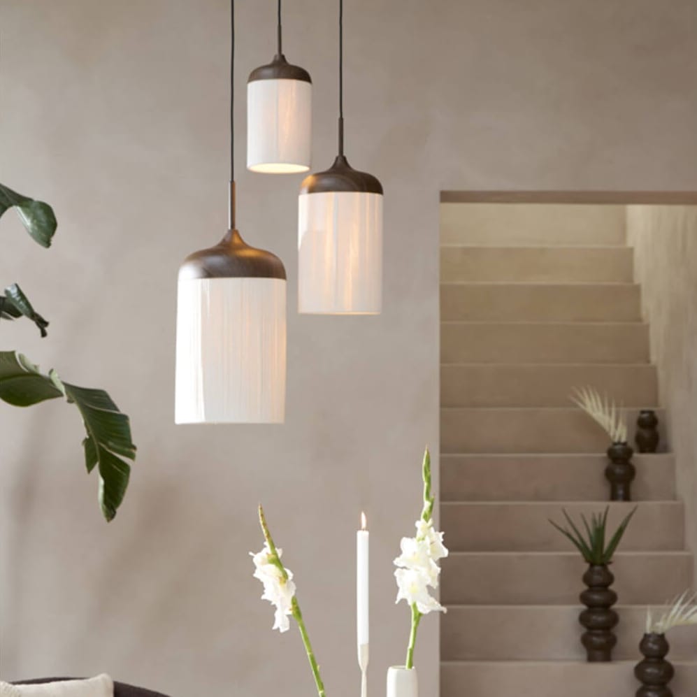 Three modern pendant lights with white cylindrical shades and dark bronze metal caps hang over a living room table. The scene includes a dark brown armchair, white vases with flowers, and a staircase in the background.