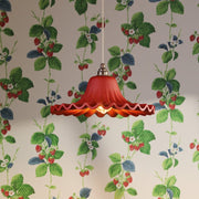 A red glass pendant light with a scalloped lower rim hangs against a cream wallpaper pattern featuring green leaves and red berries.