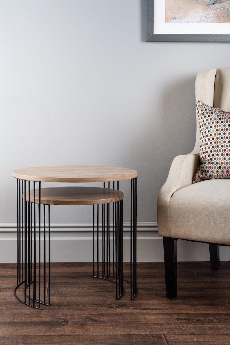 Two wooden side tables with metal legs next to a beige armchair with a patterned pillow on a wooden floor.