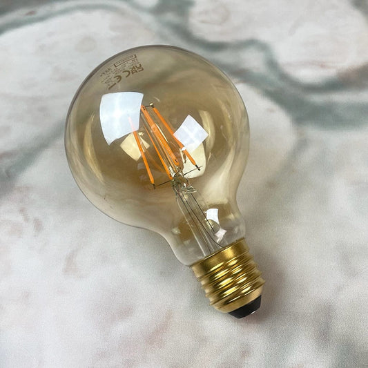 An E27 amber-colored globe LED light bulb with a dimmable feature, displayed against a textured background.