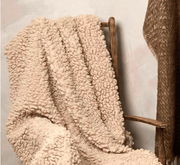 Beige sherpa blanket draped over a wooden chair against a neutral wall.