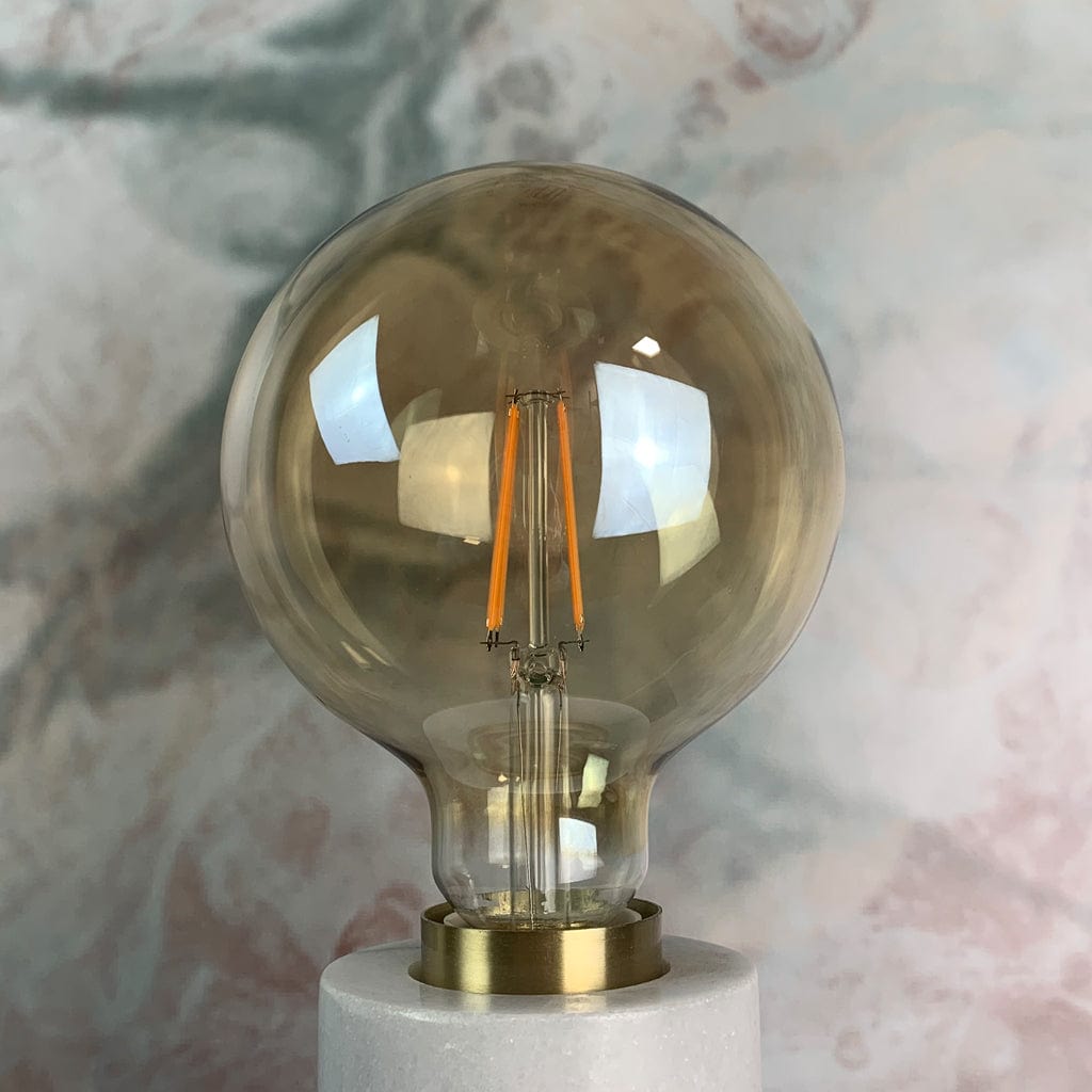 An E27 amber tinted LED globe light with a 125mm diameter and a 7.5W power rating, displayed against a marble background, in a lamp holder