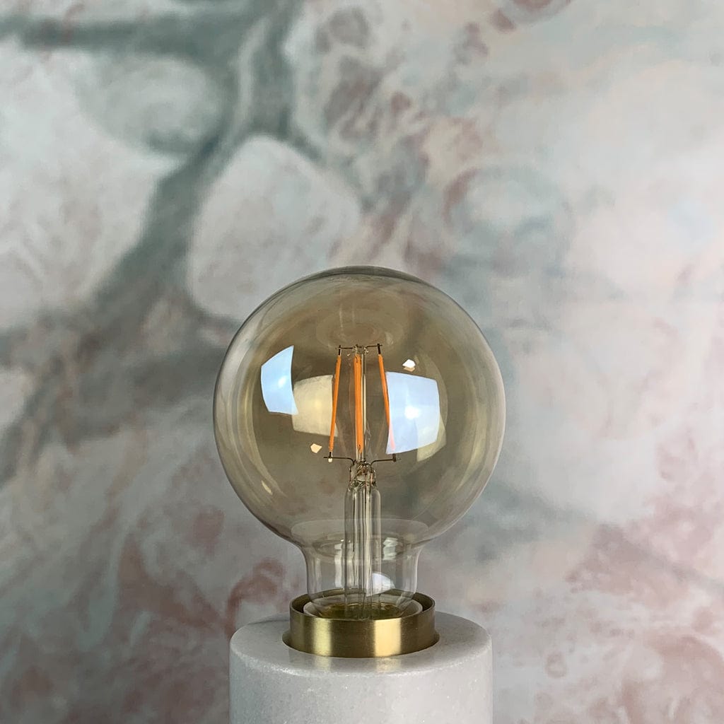 An amber tinted LED globe light bulb with a screw base, placed against a marble background, in a lamp holder