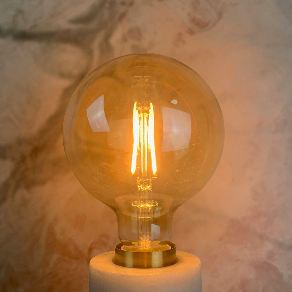An amber tinted LED globe light bulb with a screw base, placed against a marble background, lit in a lamp holder