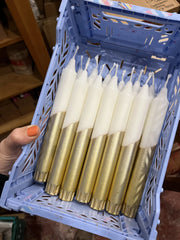 A hand with orange nail polish holds a light blue plastic crate filled with multiple taper candles. The candles have white wax tops and metallic gold wax bottoms, set against a blurry indoor background.