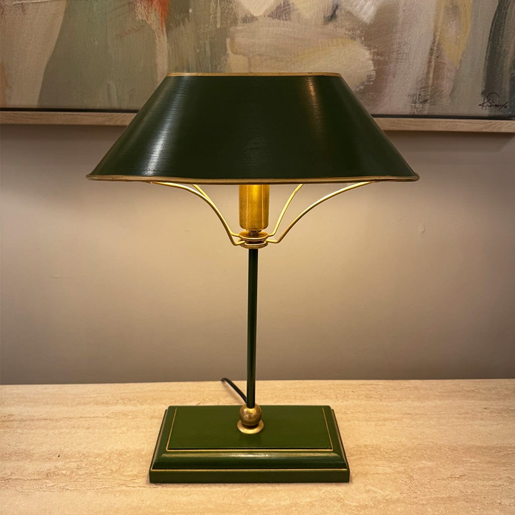 Green table lamp with gold accents on a wooden surface, with a painting in the background.