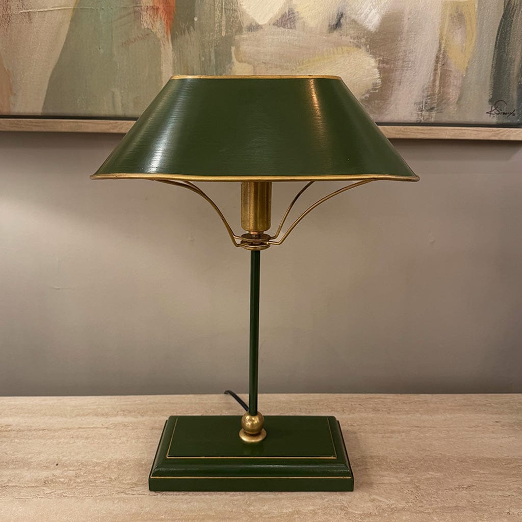 Green table lamp with gold accents on a wooden surface, with abstract art in the background.