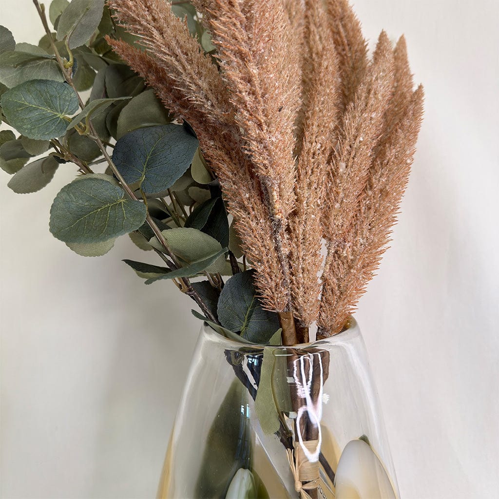 Clear glass vase with dried pampas grass and green leaves on a light background
