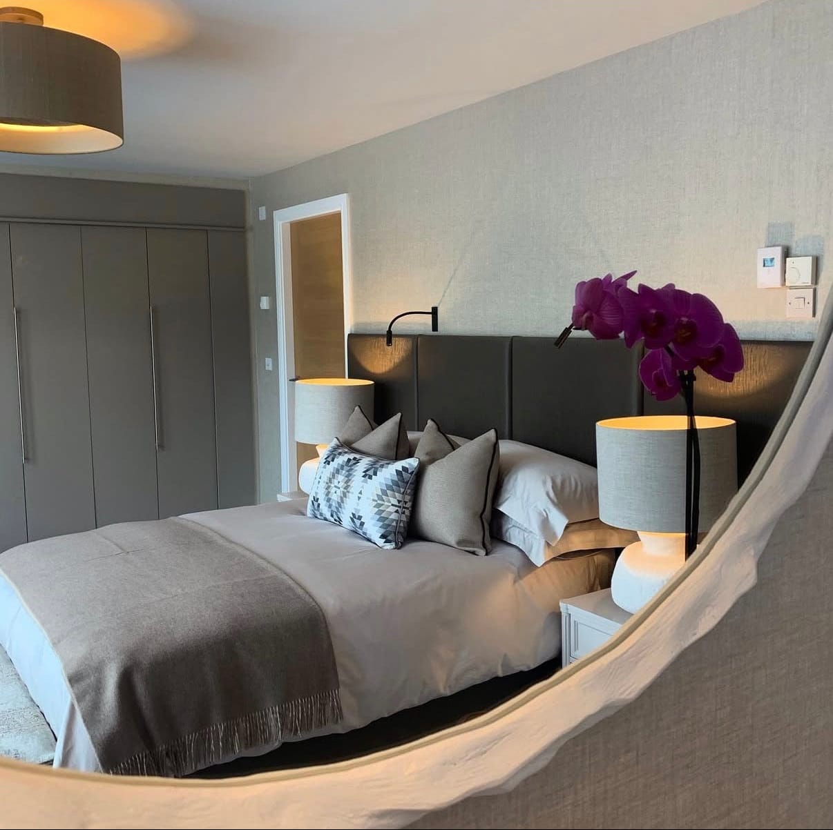 Modern bedroom with gray bedding, decorative pillows, and a purple flower arrangement.