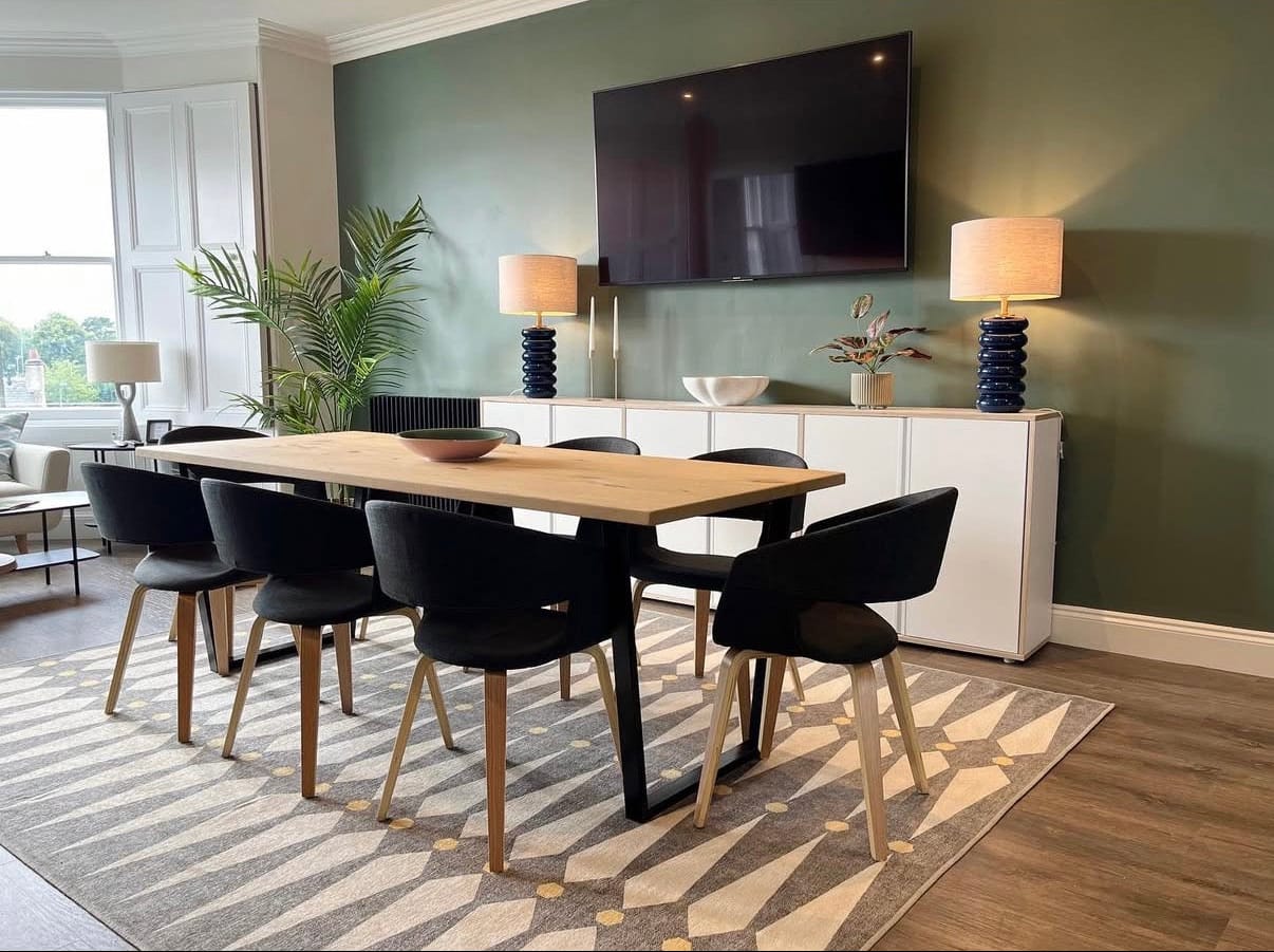 Modern dining room with a long wooden table and black chairs, featuring a TV on the wall.