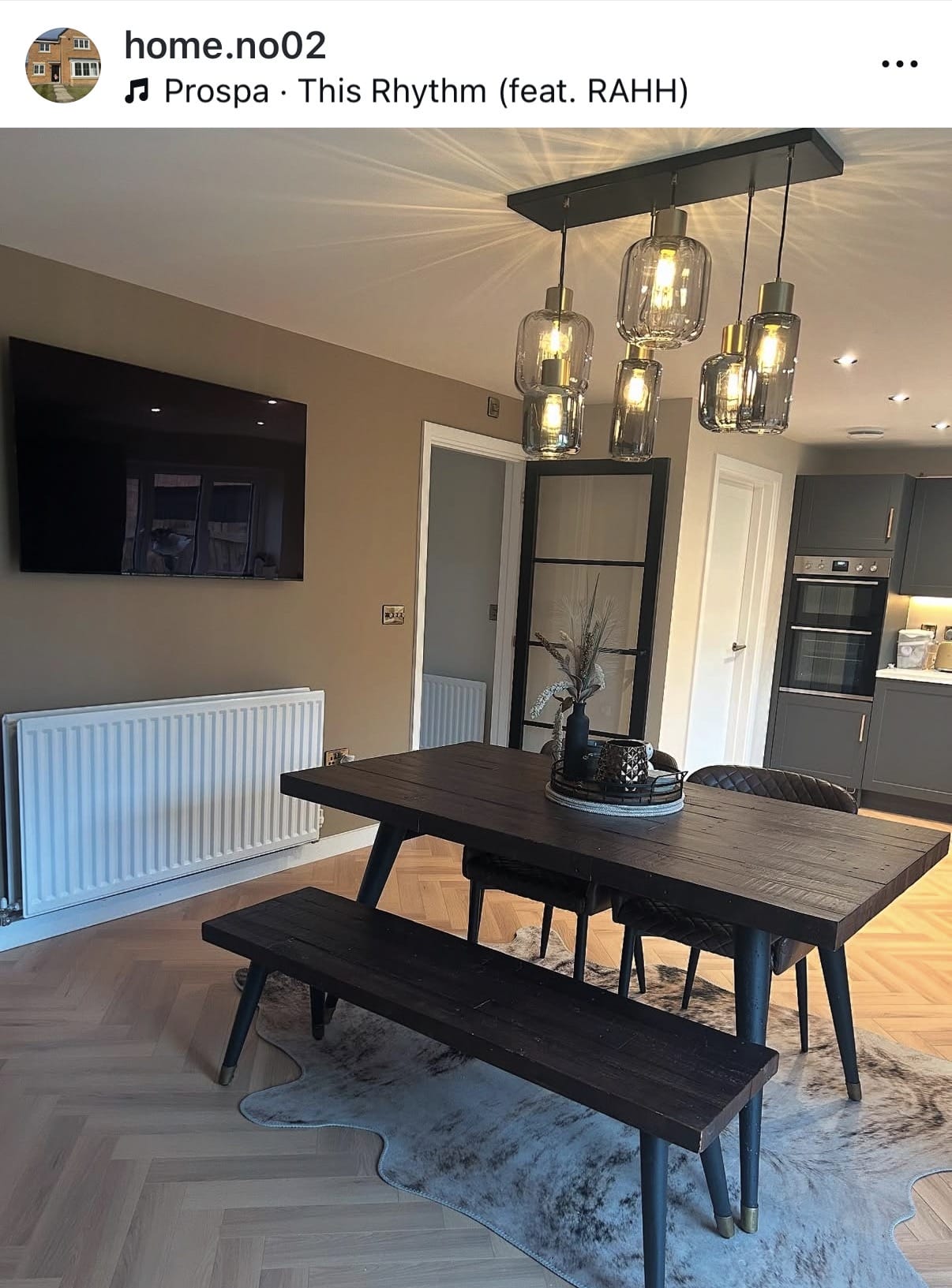 Modern dining room with a wooden table and bench, pendant lights, and a kitchen in the background.