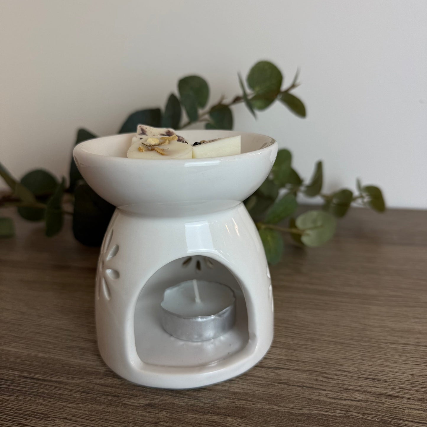 White ceramic oil burner with a candle on a wooden surface with greenery in the background