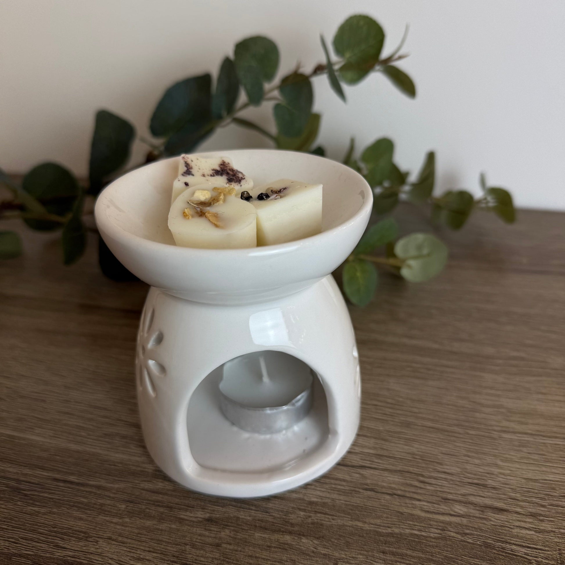 White ceramic wax melt burner with wax melts on a wooden surface with greenery in the background