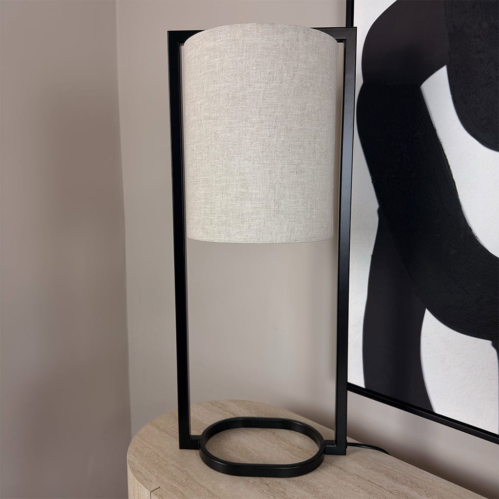 Table lamp with white shade and black base on a wooden surface, with abstract art in the background.